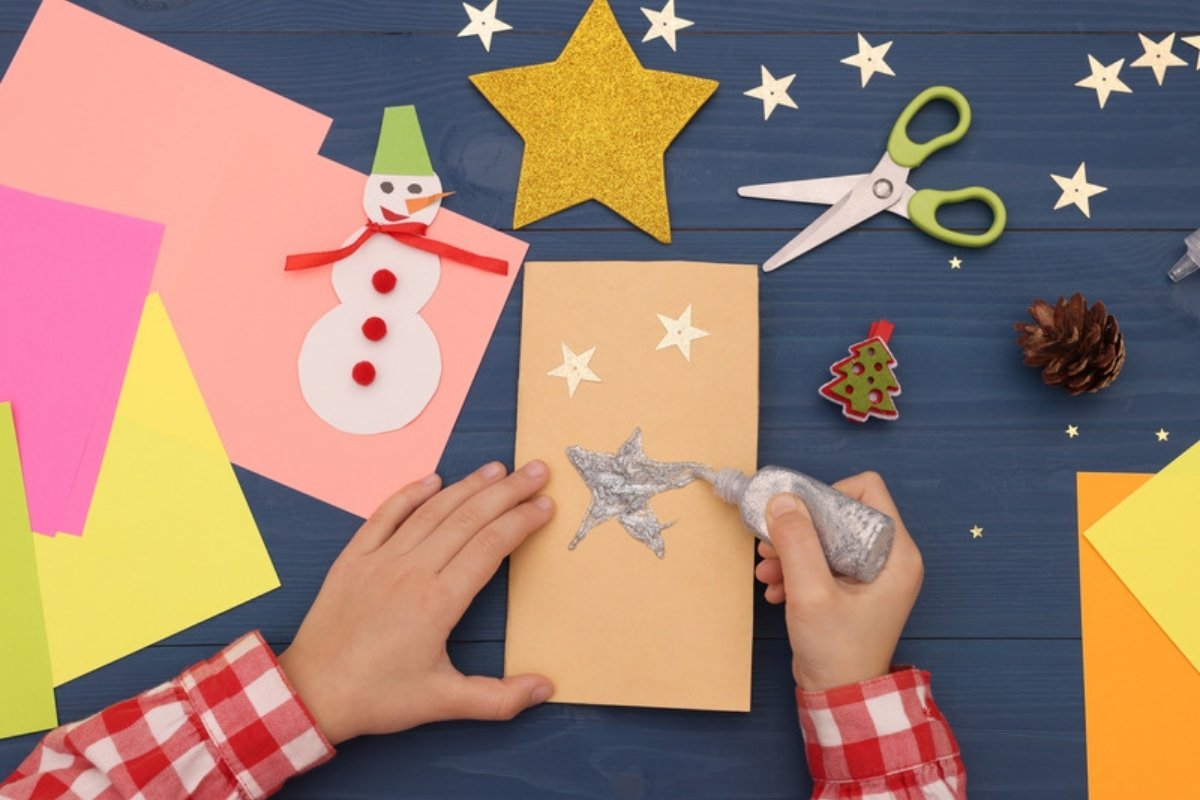 Child's hands making a paper Christmas card with a snowman, stars and glitter glue