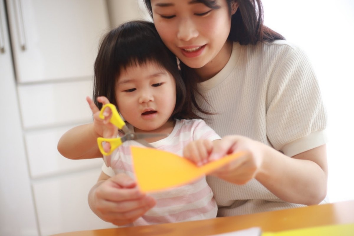 Mother helping her toddler cut yellow paper with child-safe scissors